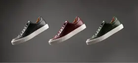 Sneakers marche famose in pelle nera, bordeaux e verde, con suola bianca, fluttuano su sfondo grigio.