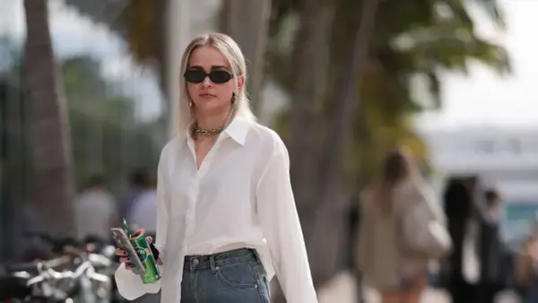 Donna con occhiali da sole, collana, camicia bianca e jeans, tiene in mano una lattina verde e un telefono.