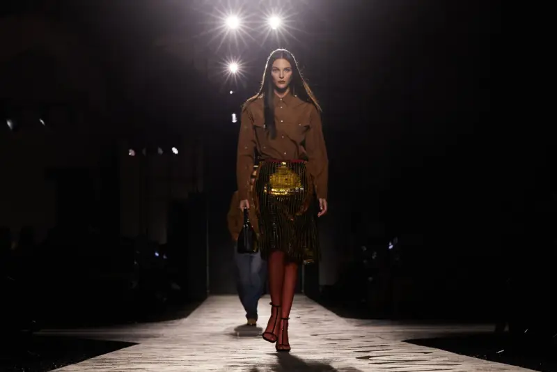 Modella su passerella, con gonna dorata e camicia marrone, un omaggio all'eleganza senza tempo di Prada, fondatore di un impero della moda.
